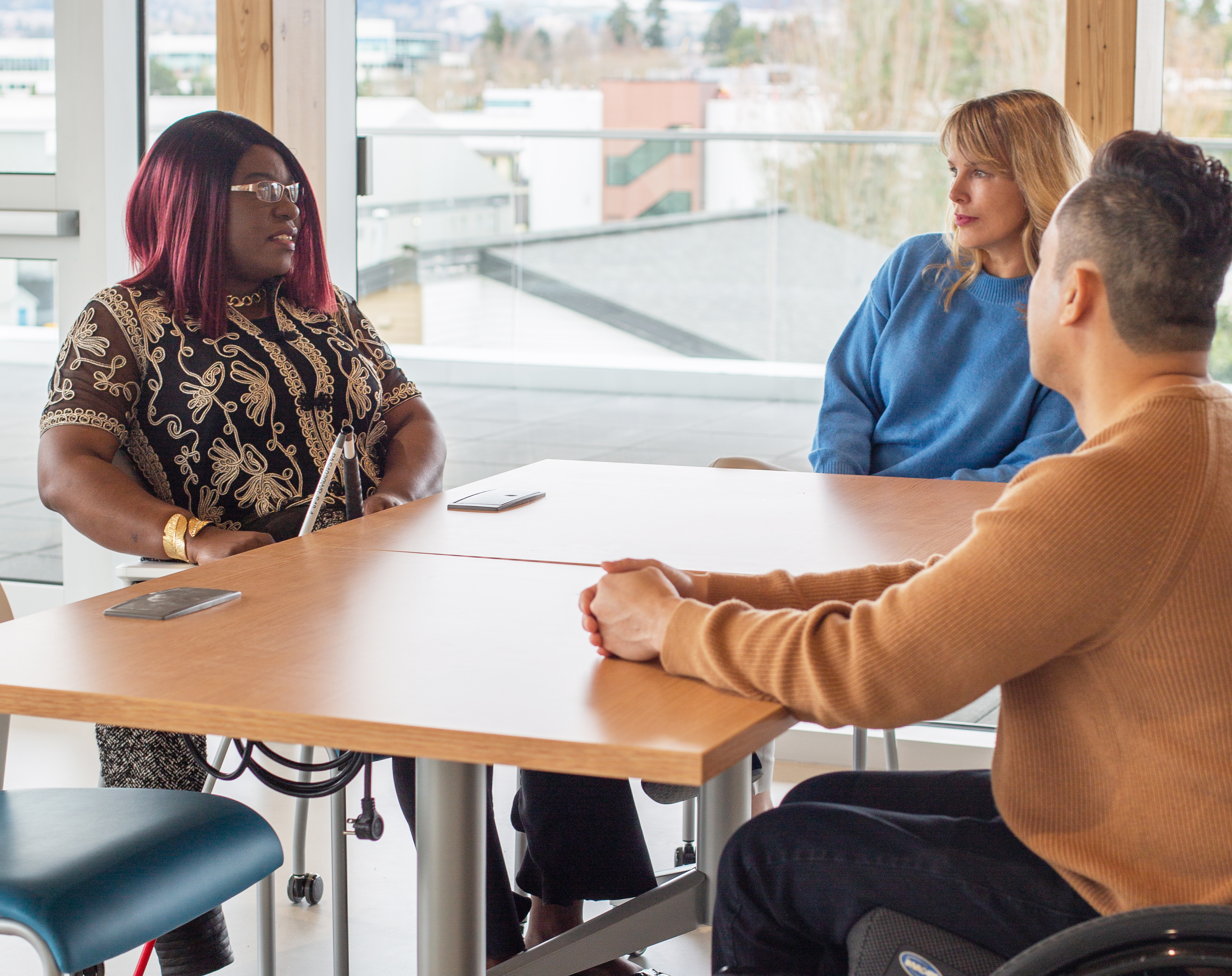 Laetitia Mfamobani and Rebecca Blissett sitting at a table with a third person who has short black hair and is wearing a tan sweater and is turned away from the camera. Laetitia is wearing a black and gold shirt and Rebecca is wearing a blue sweater.